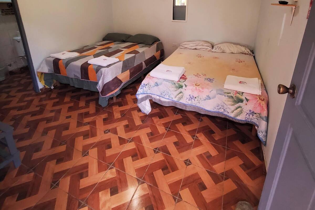 Una habitación con dos camas, suelo estampado, una lámpara de techo, equipo de aire acondicionado y una ventana pequeña.