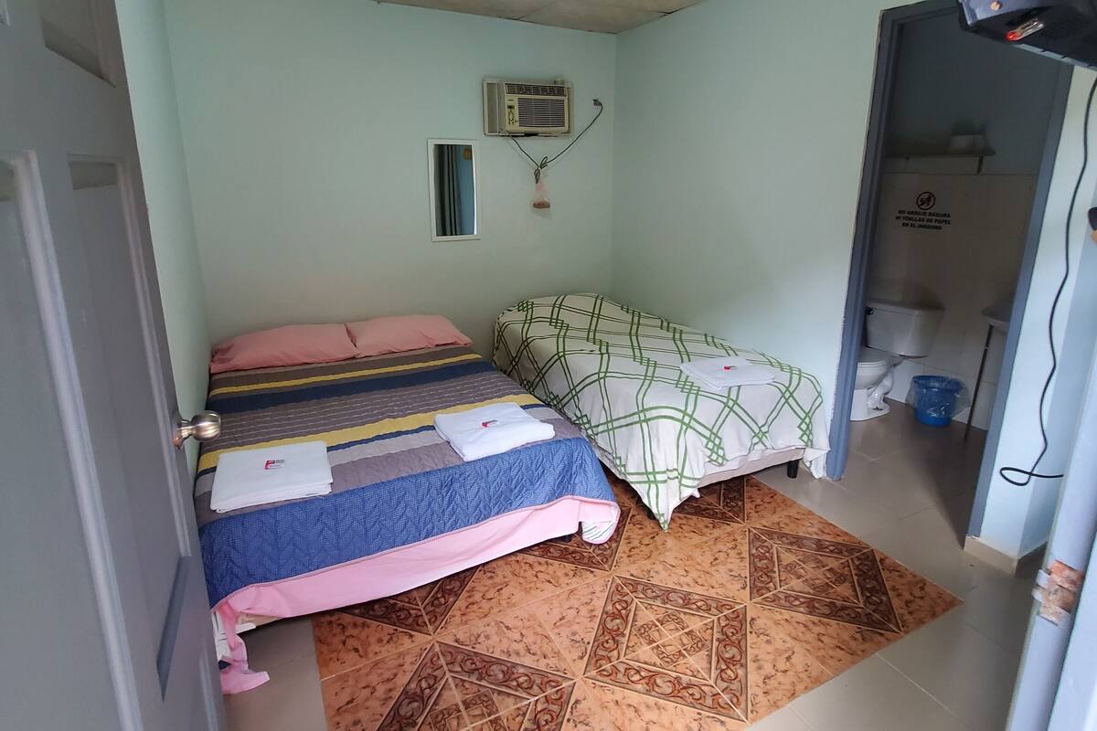 Un dormitorio con dos camas, un ventilador de techo, un equipo de aire acondicionado y una alfombra estampada.