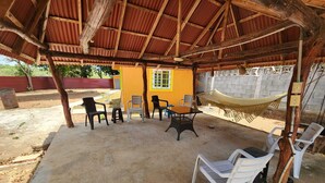 Un patio exterior con una zona de estar techada, que incluye sillas de plástico y una mesa, bajo una estructura de techo de madera.
