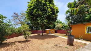 Un barrio residencial con un patio de tierra, un pequeño edificio con detalles amarillos y un árbol grande.
