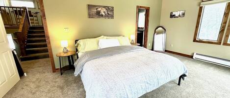 A bedroom with a large bed, a ceiling fan, a window with blinds, a mirror, and a staircase leading to an upper floor.
