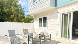 Une terrasse extérieure avec deux canapés et une table basse, une clôture blanche et une porte-fenêtre coulissante donnant sur un espace intérieur.