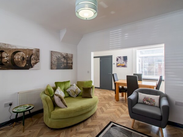 A modern living room with a green sofa, a grey armchair, and a dining area with a wooden table and chairs.
