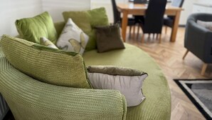 A green sofa with multiple cushions in a living room with a dining area in the background.
