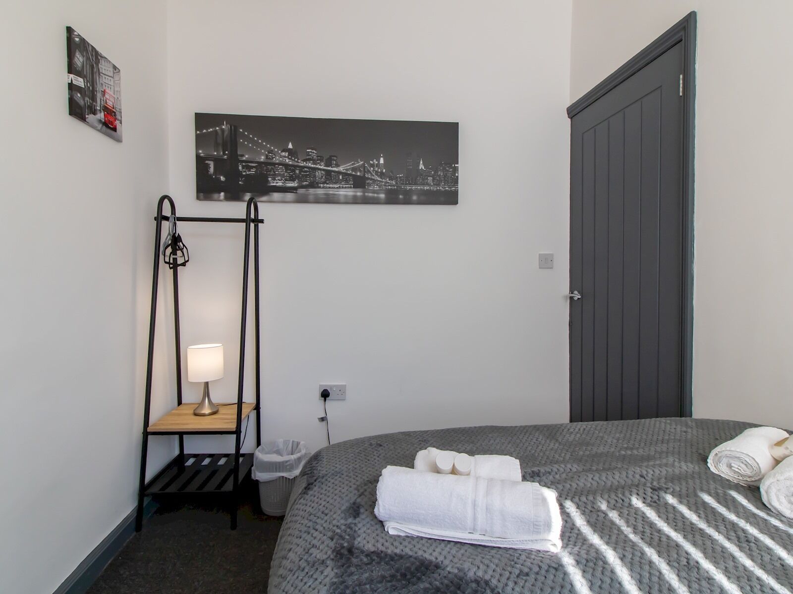 A hotel room with a bed, a nightstand, a lamp, a picture of a cityscape, and a door.