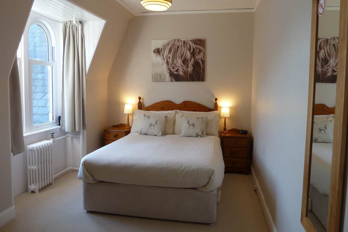 A bedroom with a bed, two bedside tables, a nightstand, a radiator, a window with curtains, a ceiling light, and a wall-mounted picture.