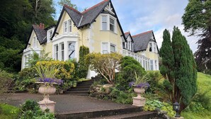 A large, two-story house with a steeply pitched roof, multiple windows, and a well-maintained garden with steps leading to the entrance. - Camus House (Onich)