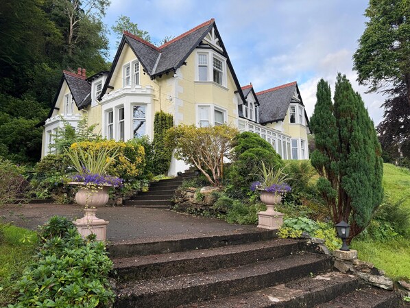 A large, two-story house with a steeply pitched roof, multiple windows, and a well-maintained garden with steps leading to the entrance. - Camus House (Onich)