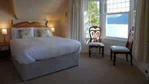 A bedroom with a large bed, a chair, a small table, and a window with a view of a body of water and mountains. - Camus House (Onich)