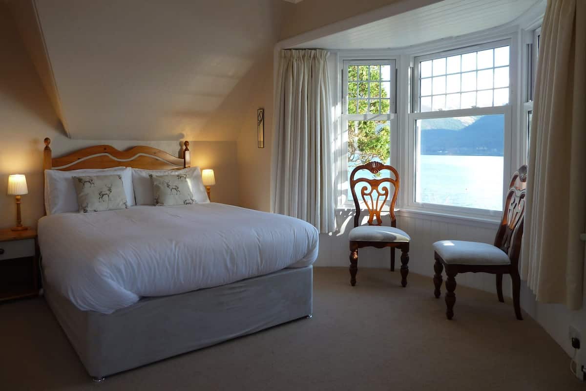 A bedroom with a large bed, a chair, a small table, and a window with a view of a body of water and mountains.