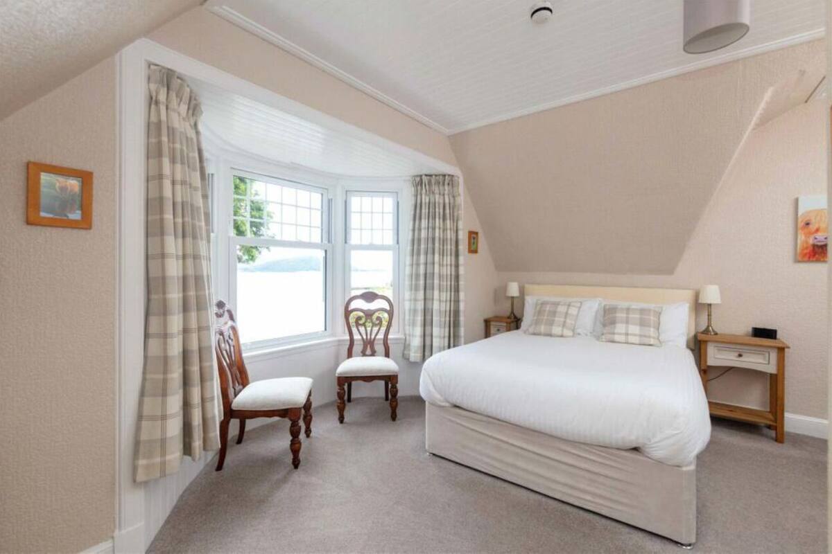 A bedroom with a large bed, two chairs, a nightstand, and a window with curtains.