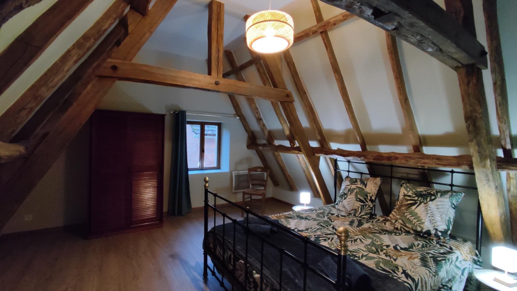 Ein Schlafzimmer mit schrägen Wänden, Holzbalken, einem Bett mit floralem Bettzeug, einem Sessel und einem Fenster mit Vorhängen.
