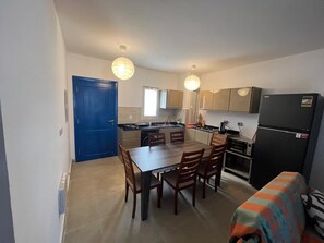 Een kleine keuken met een eettafel, stoelen, een blauwe deur en een koelkast.