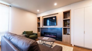 Una sala de estar moderna con un sofá de cuero, una mesita redonda pequeña, un televisor de pantalla plana montado en una pared de madera, una chimenea y estanterías empotradas.