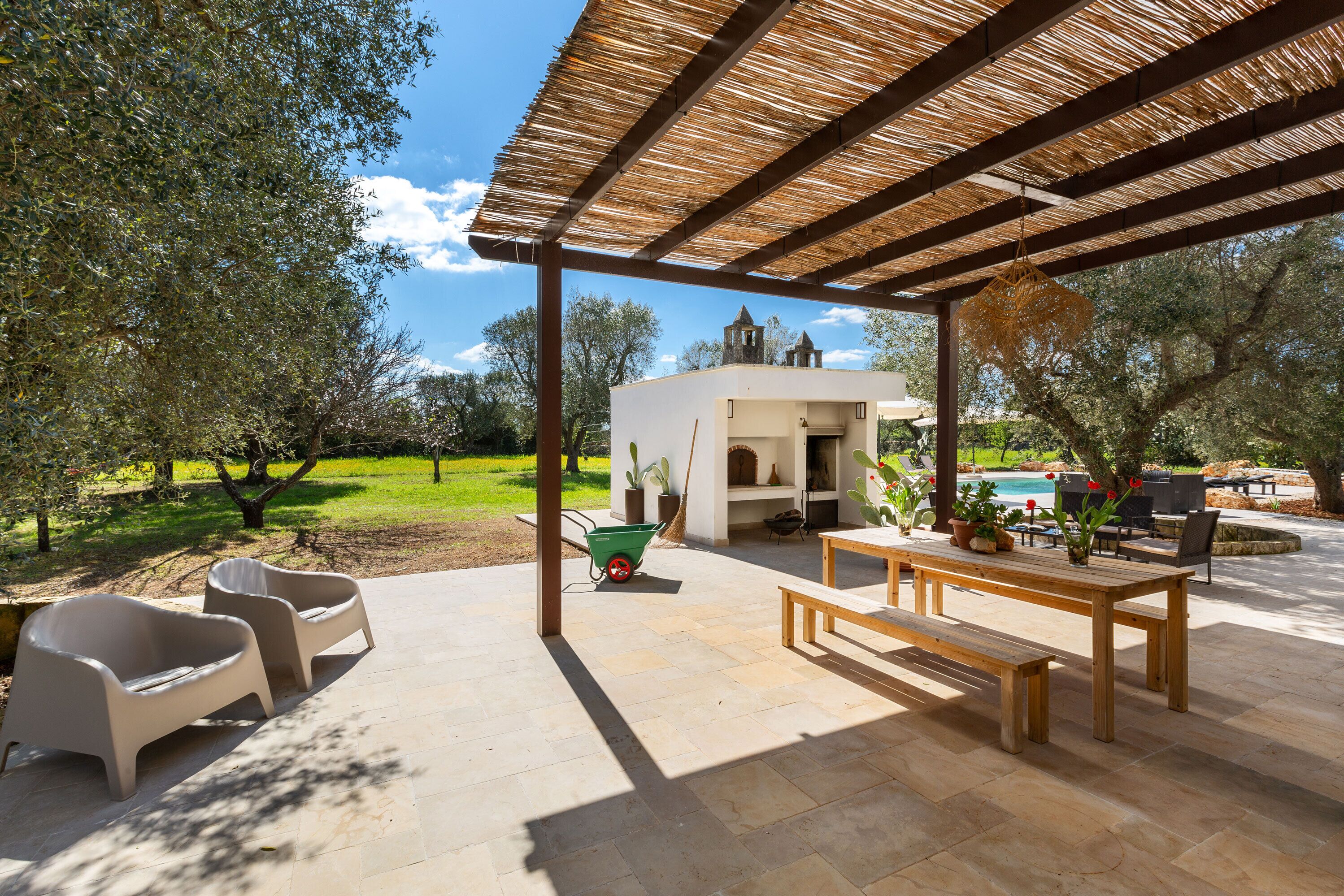 An outdoor patio with a pergola, seating, a table, and a view of a pool and garden.
