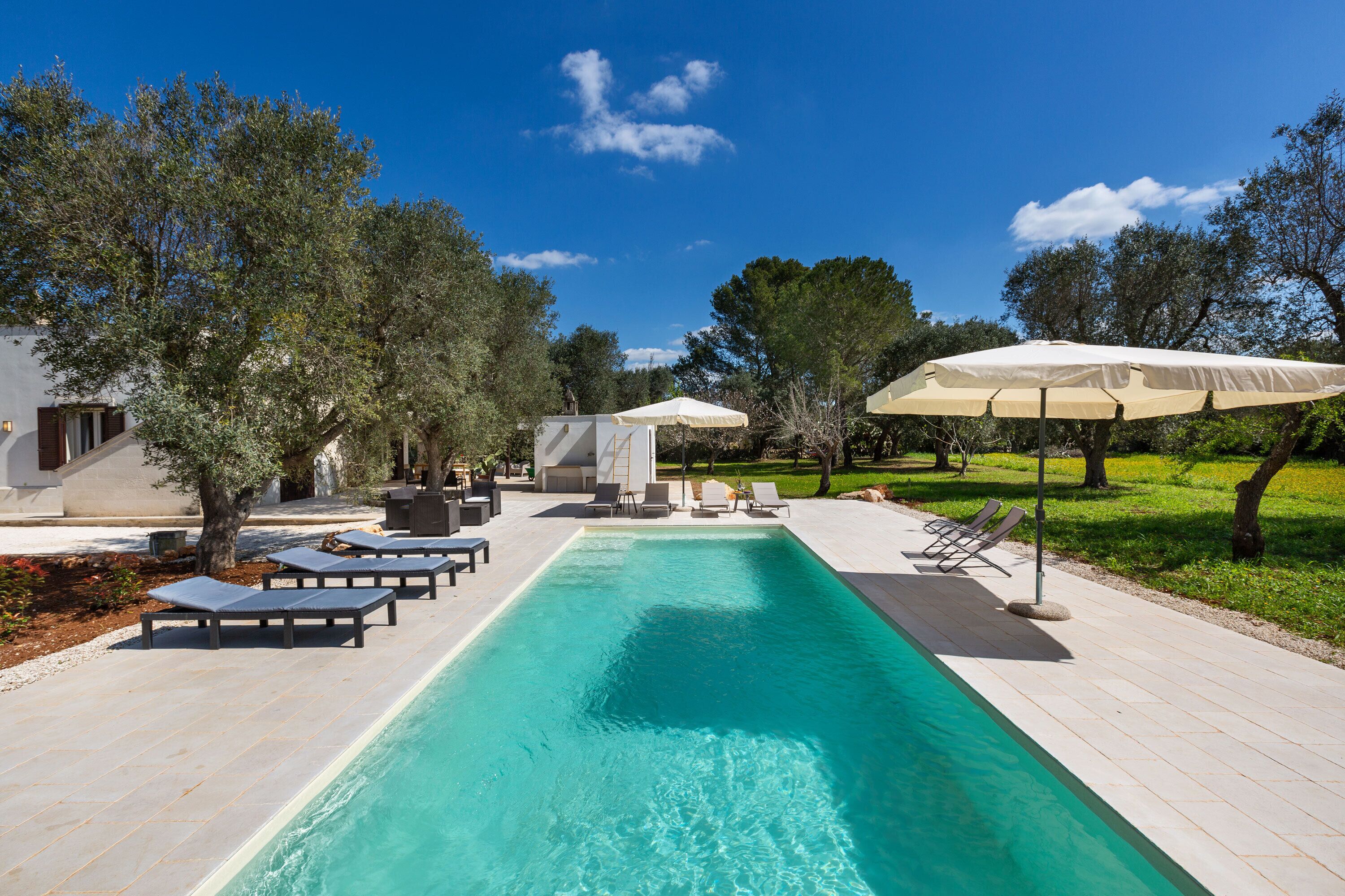 Una piscina con lettini e ombrelloni, circondata da alberi e un cielo sereno.