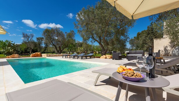Zona de piscina con sillas de descanso, una mesa de comedor con comida y bebidas, y un cielo azul despejado.
