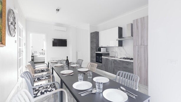 Un comedor moderno con una larga mesa dispuesta para cuatro, sillas blancas y una cocina al fondo.