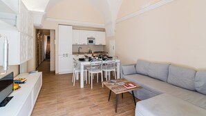 A modern living room with a grey sofa, a coffee table, and a TV. There is a kitchen area with white cabinets and a dining table with chairs. - Casa Margot by BarbarHouse (Campomarino)