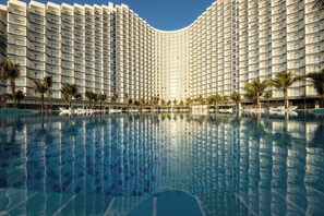 Un hotel grande con fachada curva, una piscina central y palmeras alrededor del área de la piscina.