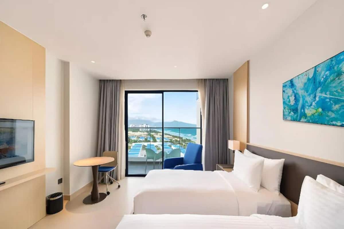 A hotel room with two beds, a TV, a dining table, and a view of a cityscape.