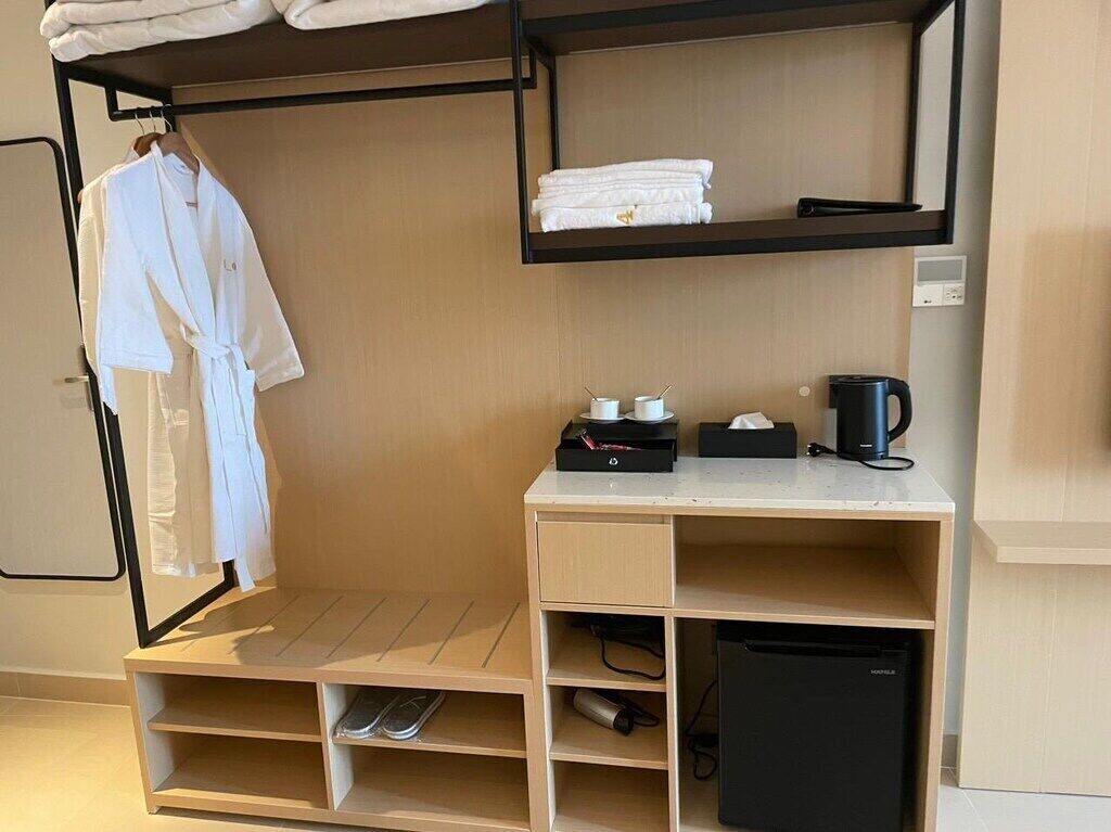 Habitación de hotel pequeña con cama, escritorio con cafetera, refrigerador negro y estante con toallas dobladas.