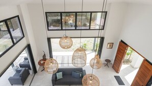 Ein geräumiger Wohnbereich mit grauem Sofa, mehreren hängenden Rattan-Lampen und großen Fenstern mit Blick ins Freie.