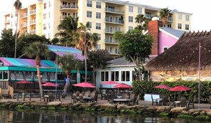 A waterfront restaurant with outdoor seating, a pool, and a building with balconies in the background.