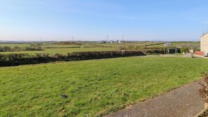 A rural landscape with a grassy field, a stone wall, and a building with a paved path leading to it. - 11 Penygroes (Holyhead)