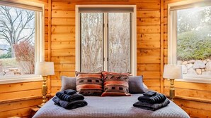A wooden cabin room with a bed, two windows, and a view of trees and rocks. - Pikes Peak Place: Relaxing Oasis w/Mtn Views (Colorado Springs)