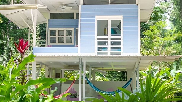 Una casa de dos pisos con un balcón, rodeada de abundante vegetación.
