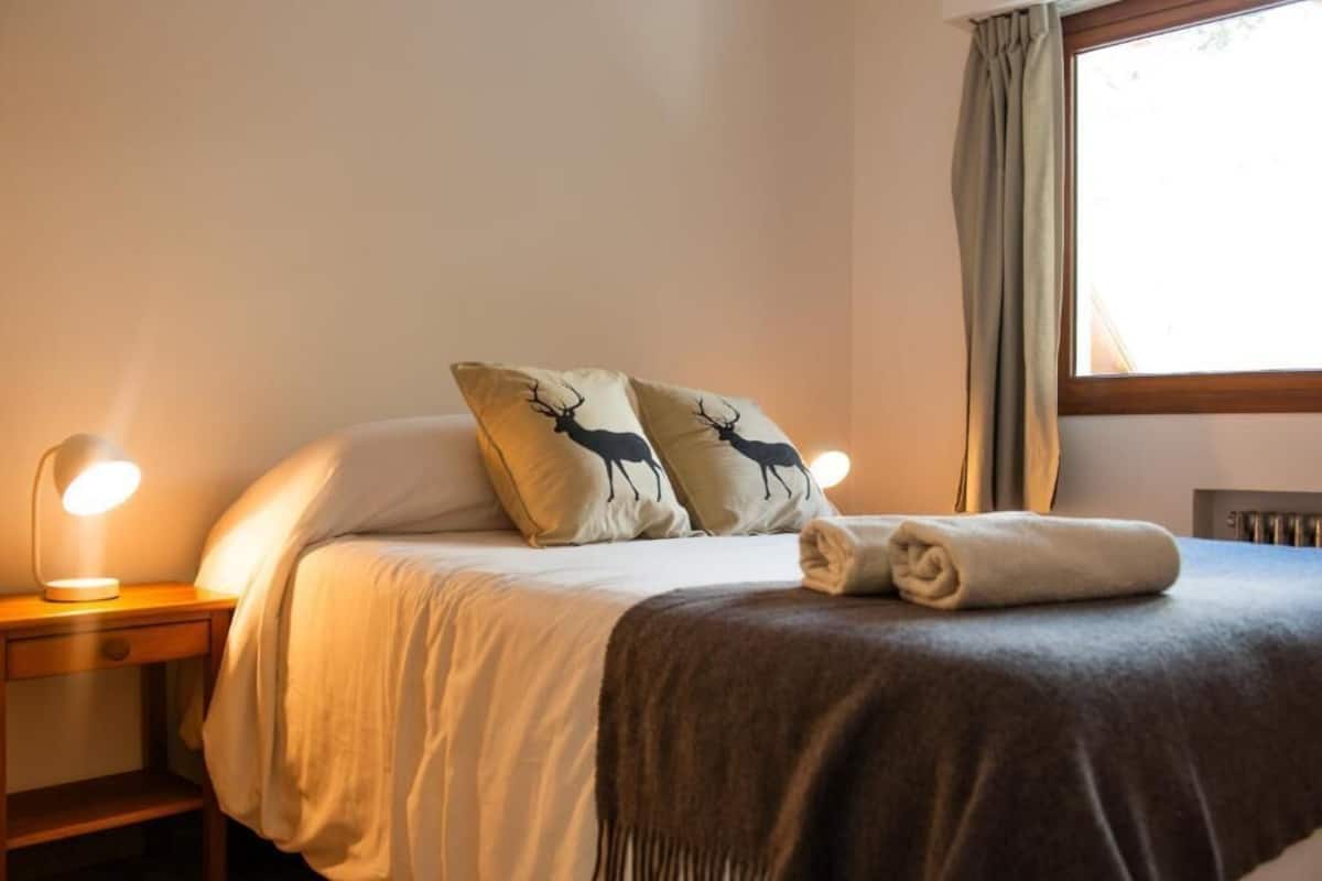 Una cama bien hecha con dos almohadas con siluetas de ciervos, una toalla doblada y una lámpara de noche sobre una mesita de madera.