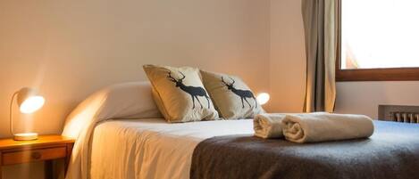 Una cama bien hecha con dos almohadas con siluetas de ciervos, una toalla doblada y una lámpara de noche sobre una mesita de madera.