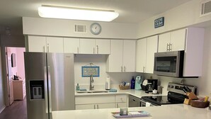 A modern kitchen with white cabinets, a stainless steel refrigerator, a microwave, and a coffee maker. There are two stools at the island and a trash can. - 1 bedroom on Okaloosa Island FtWalton1 minute walk to the beach. Clean,spacious. (Fort Walton Beach)