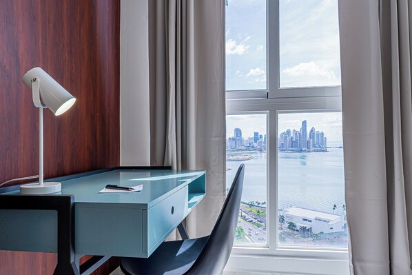 A modern office space with a desk, chair, and lamp. A large window offers a view of a city skyline and water.