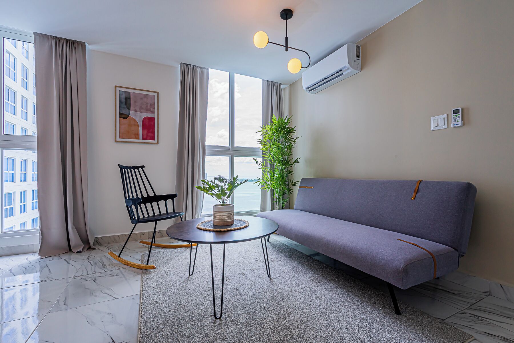 A modern living room with a grey sofa, a round table with a plant, a black chair, and a large window with curtains.