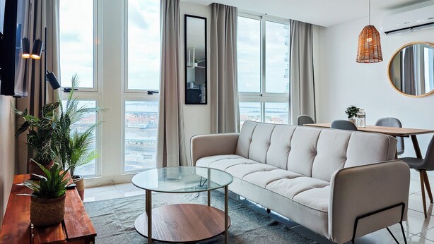 A modern living room with a sofa, a glass coffee table, a dining table with chairs, and large windows offering a city view.