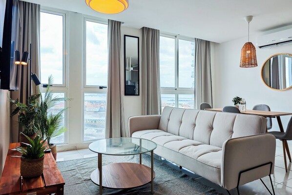 A modern living room with a sofa, a glass coffee table, a dining table with chairs, and large windows offering a city view.