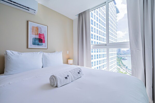 A hotel room with a large bed, a framed abstract painting, a rolled towel, and a view of tall buildings and a river through the window.