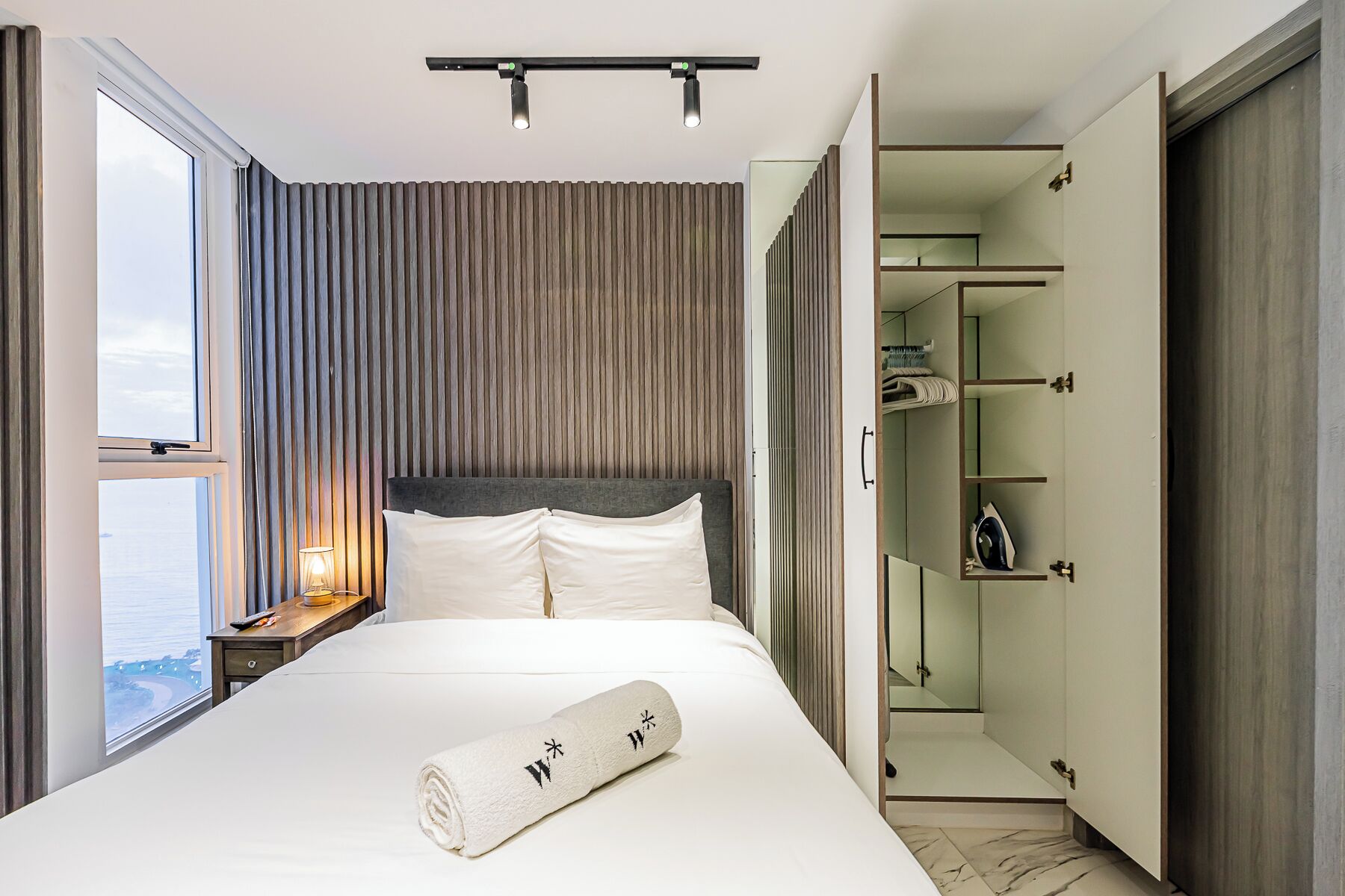 A modern hotel room with a large bed, a nightstand, a bedside lamp, a window with curtains, a built-in wardrobe, and a view of the sea.