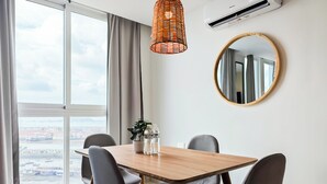 A modern room with a wooden dining table, four chairs, a hanging light fixture, a round mirror, and a large window with a view of a cityscape.