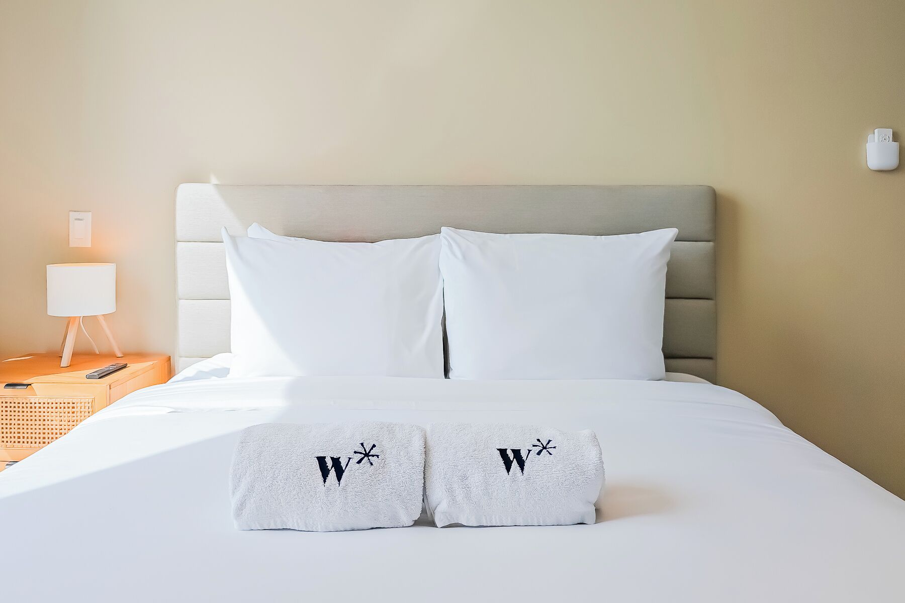 Ein Hotelzimmer mit einem Bett, zwei Kissen, einem Nachttisch mit Lampe und zwei Handtüchern mit eingestickten Initialen.