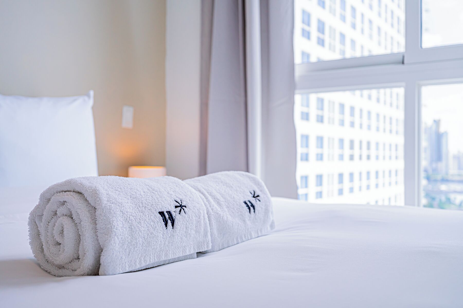 Asciugamano bianco ben disposto con scritta ricamata su un letto in una camera d'albergo con vista sulla città.