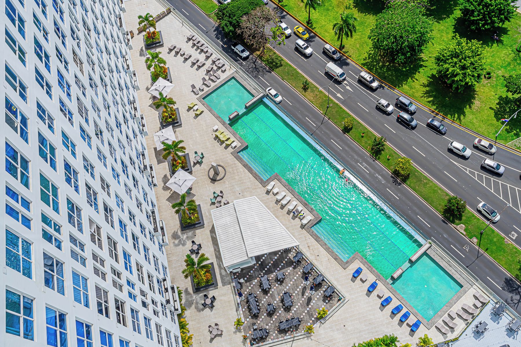 Un hôtel de grande hauteur doté d’une piscine sur le toit, de plusieurs piscines et situé près d’une route où circulent des véhicules.