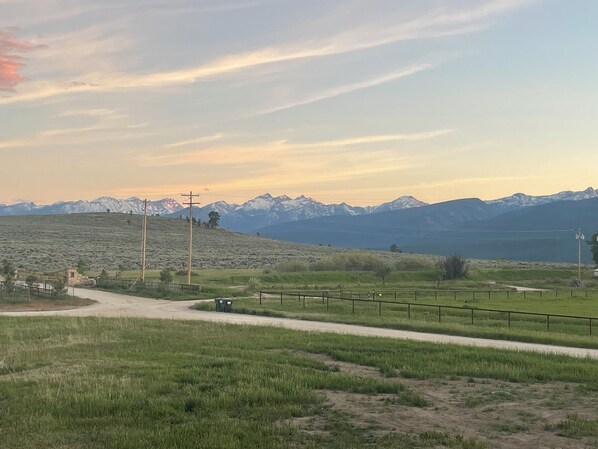 A rural landscape with a dirt road, green fields, and distant snow-capped mountains under a clear sky.