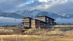 A two-story house with a flat roof and a balcony, set against a backdrop of mountains and a cloudy sky.