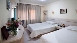 A hotel room with two beds, a desk with a vase of flowers, a bedside table with a lamp, and a window with curtains. - Jinqilin Hostel (Shanghai Waigaoqiao Free Trade Zone) (Shanghai)