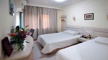 Quarto de hotel com duas camas, uma escrivaninha com um vaso de flores, uma mesa de cabeceira com abajur e uma janela com cortinas.