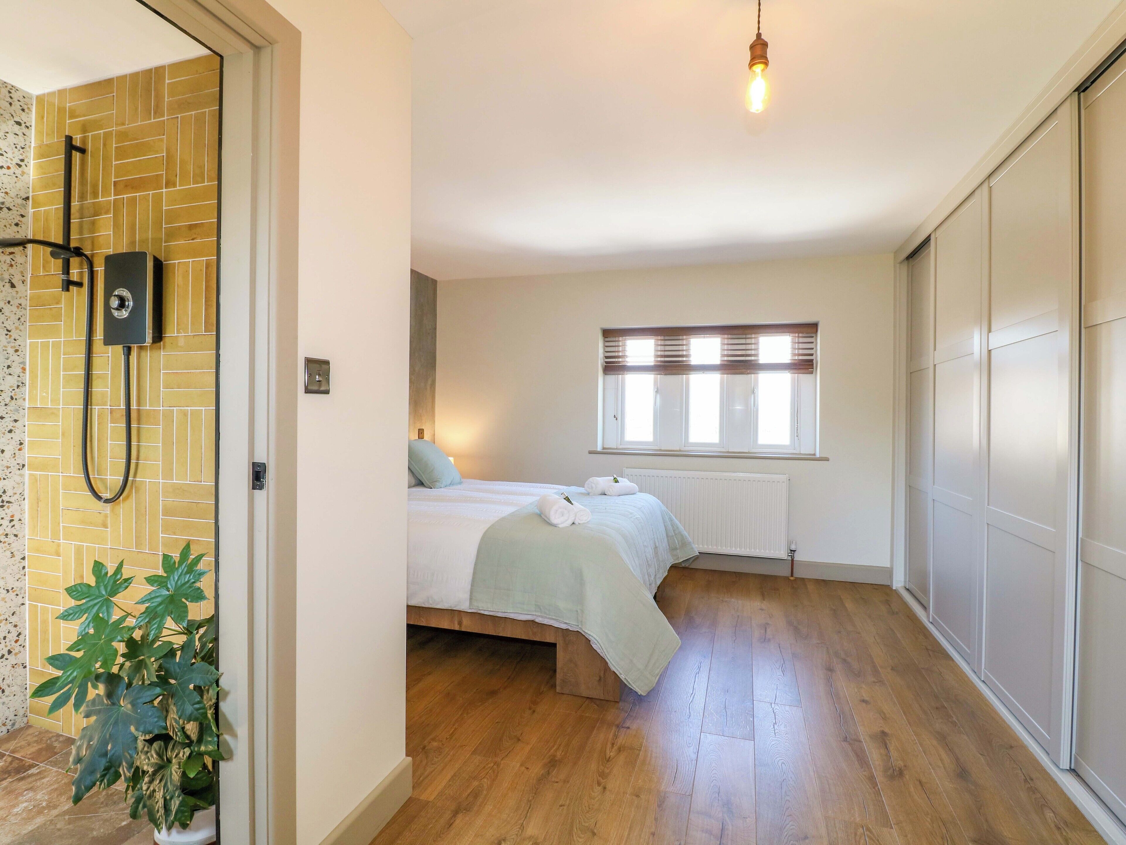 Una camera da letto con pavimento in legno, un letto con copriletto verde, una finestra con persiane e un angolo doccia con pannello di controllo a parete.