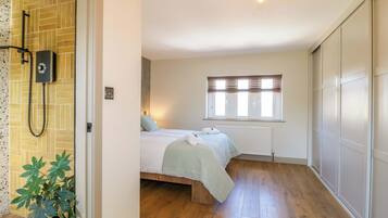 A bedroom with a wooden floor, a bed with a green bedspread, a window with blinds, and a shower area with a wall-mounted control panel.